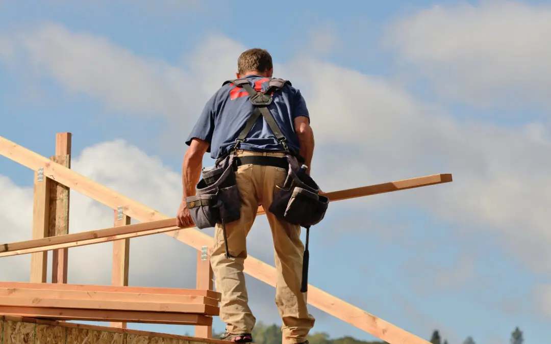 A construction worker wearing a tool belt stands on a wooden structure, holding a beam, with a blue sky and clouds in the background.