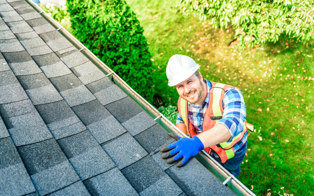 A construction worker in a hard hat, safety vest, and gloves stands on a ladder next to a shingled roof, cleaning or inspecting the rain gutter as part of regular roof maintenance recommended for homeowners.