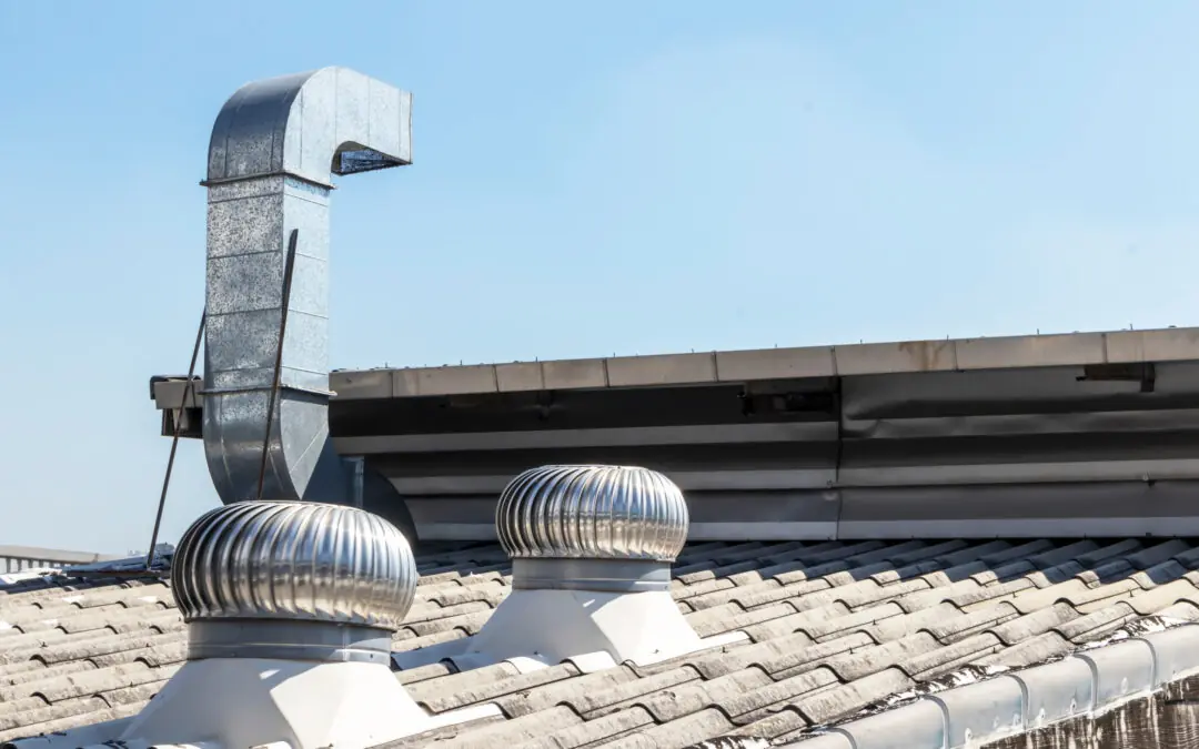 Metal ventilation ducts and turbine vents installed on the tiled roof of an industrial building, with a clear blue sky in the background.