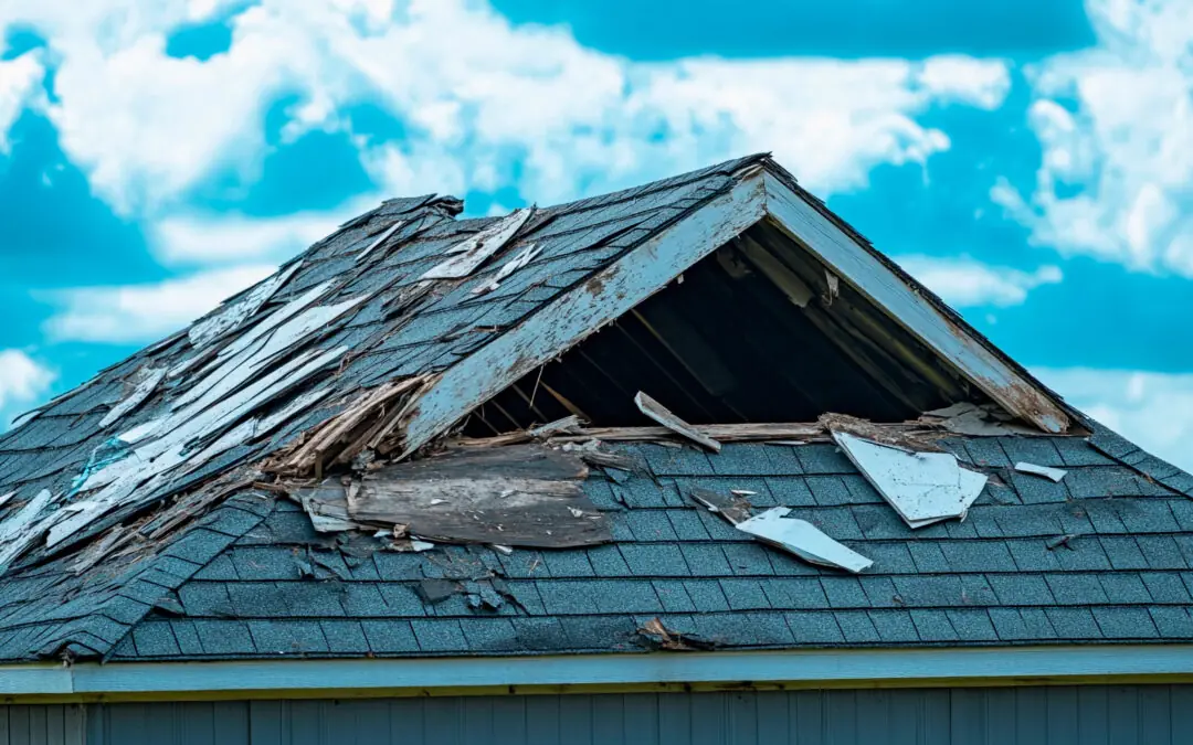 A house roof with significant damage, including missing shingles, exposed wood, and broken sections, under a partly cloudy sky.