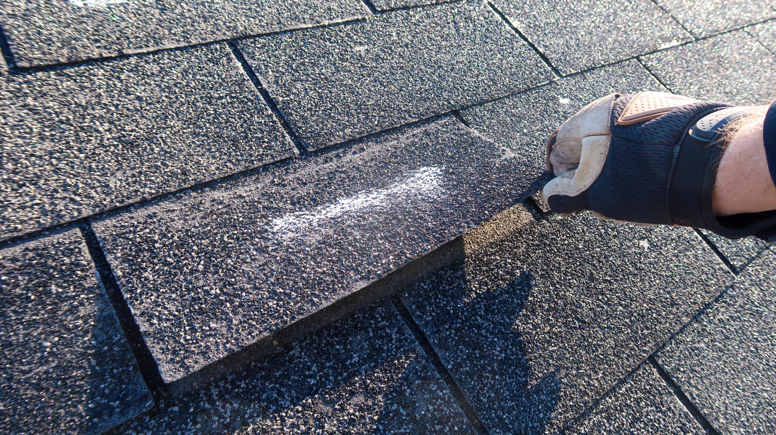 A gloved hand lifts an asphalt roof shingle marked with a white chalk line, checking for storm damage and spot roof issues.
