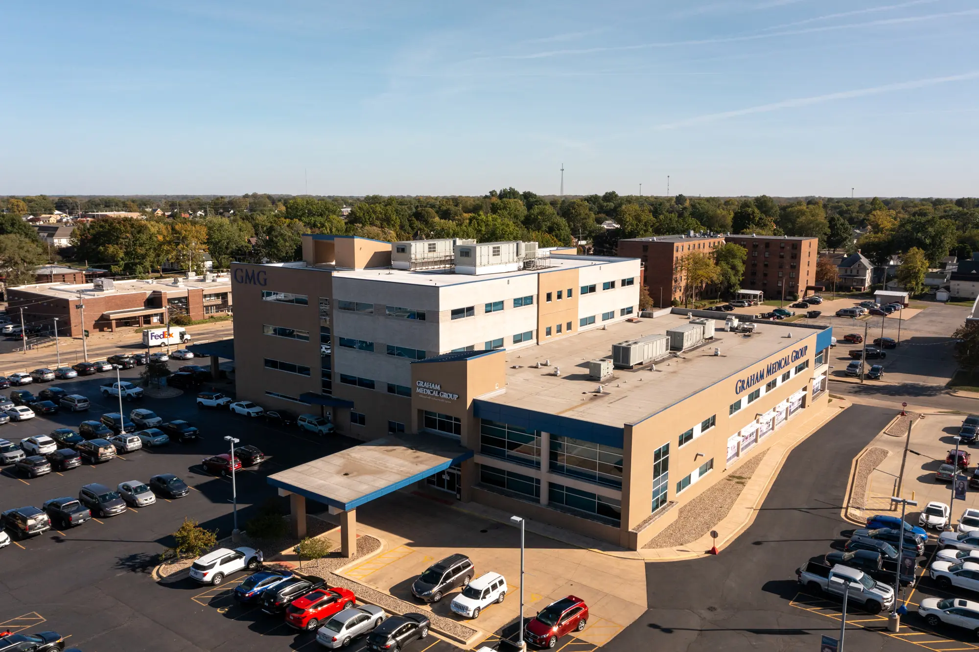 Aerial view of a hospital building with a well-maintained roof and expansive parking lot, surrounded by trees and other buildings under a clear sky.