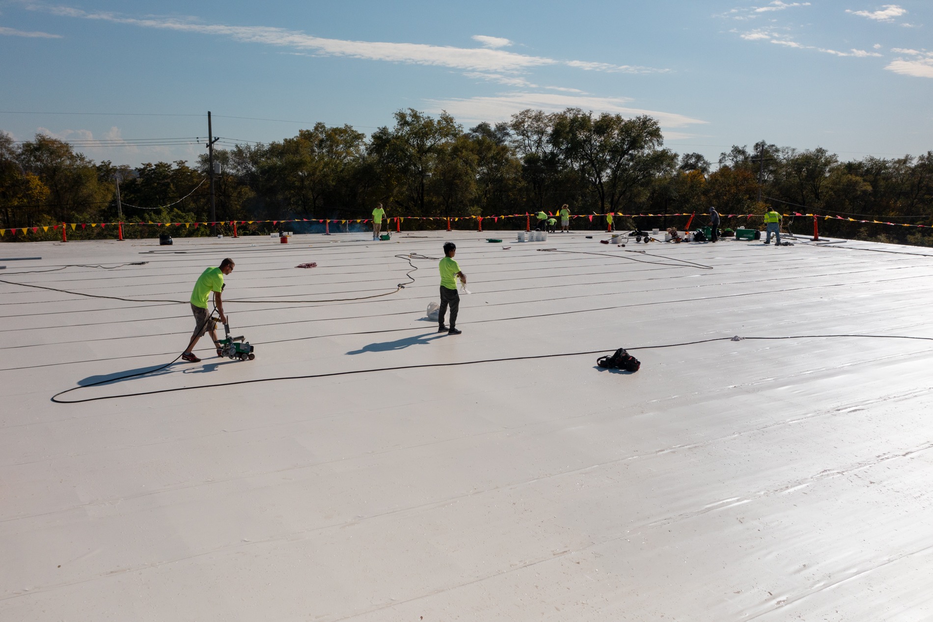 Workers in neon shirts install material on a large flat white rooftop, with tools and cables spread across the surface and trees in the background.