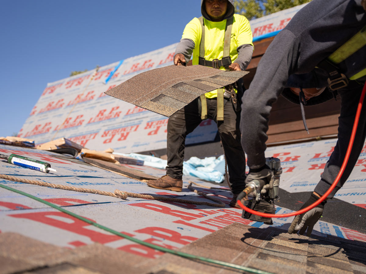 Two workers in safety gear install shingles on a house roof under clear skies, using a nail gun and construction materials.