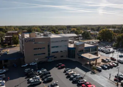 Aerial view of a hospital building, its roof meticulously maintained by Holthaus Roofing Services. The parking lot is full of cars, with signs reading "GMG" and "Patient Tower Entrance." The surrounding area includes trees and other buildings.