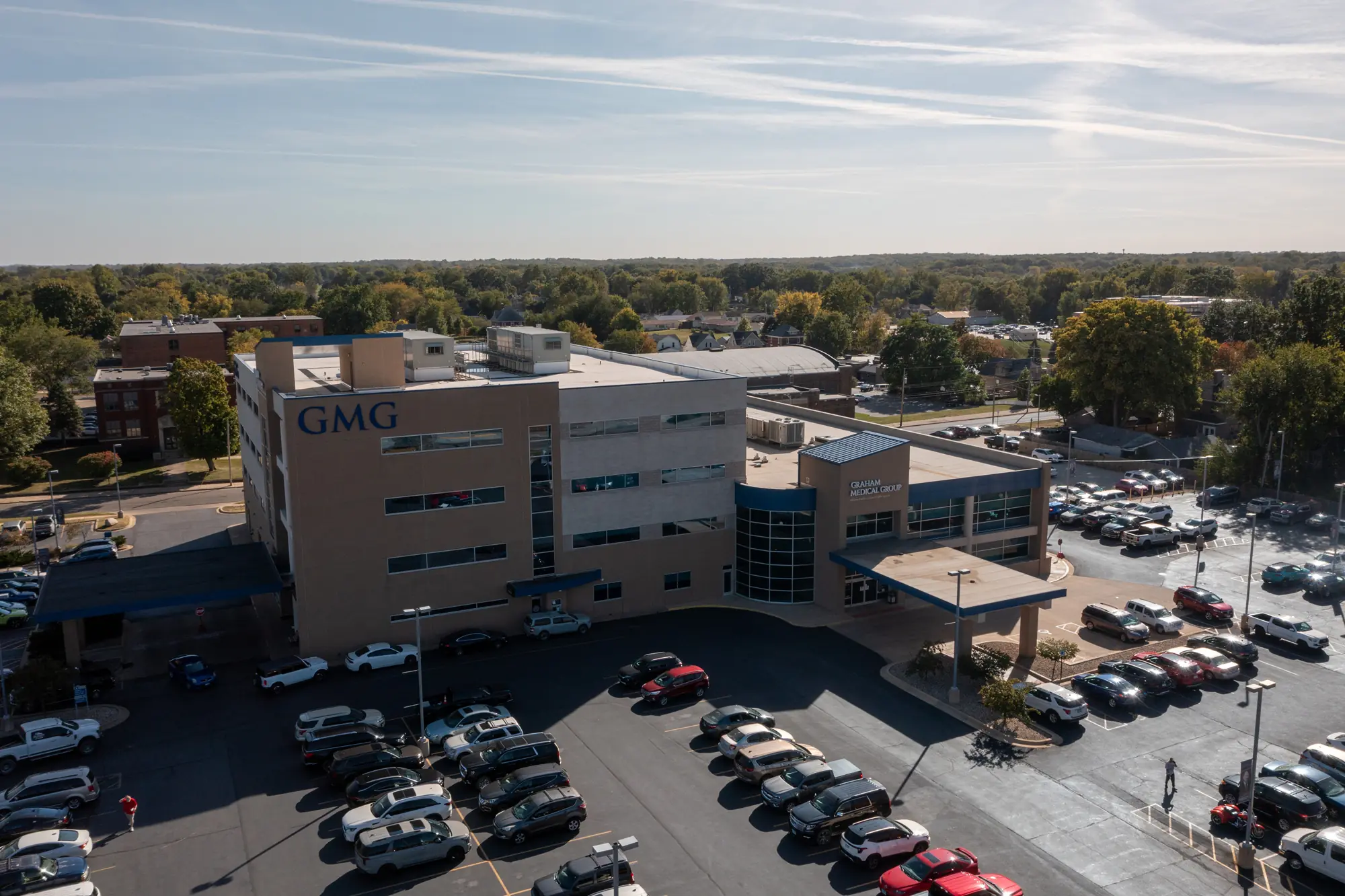 Aerial view of a hospital building, its roof meticulously maintained by Holthaus Roofing Services. The parking lot is full of cars, with signs reading "GMG" and "Patient Tower Entrance." The surrounding area includes trees and other buildings.