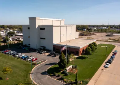 An aerial view of the Holthaus industrial building showcases a spacious parking lot filled with cars, all surrounded by lush grassy areas and a neatly paved road. The day is clear, highlighting the structure's craftsmanship and hinting at reliable roofing services.