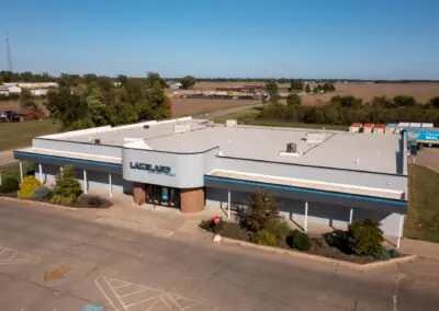 Aerial view of a large, single-story commercial building with a white roof by Holthaus Roofing Services and the sign "Lakeland" on the front. It's surrounded by a parking area and greenery, set against a rural landscape.