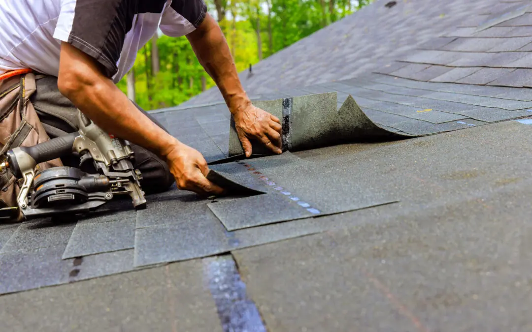 A person installs asphalt shingles on a roof using a nail gun and hands, highlighting essential roof repair as part of meeting your roof needs, with a wooded background visible.