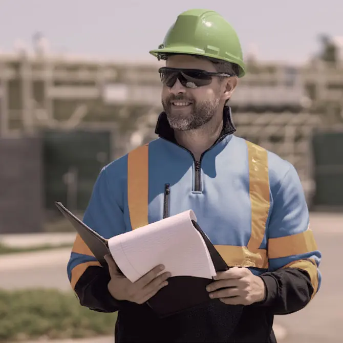 A construction worker wearing a green hard hat and safety vest holds a clipboard and notebook outdoors at a work site.