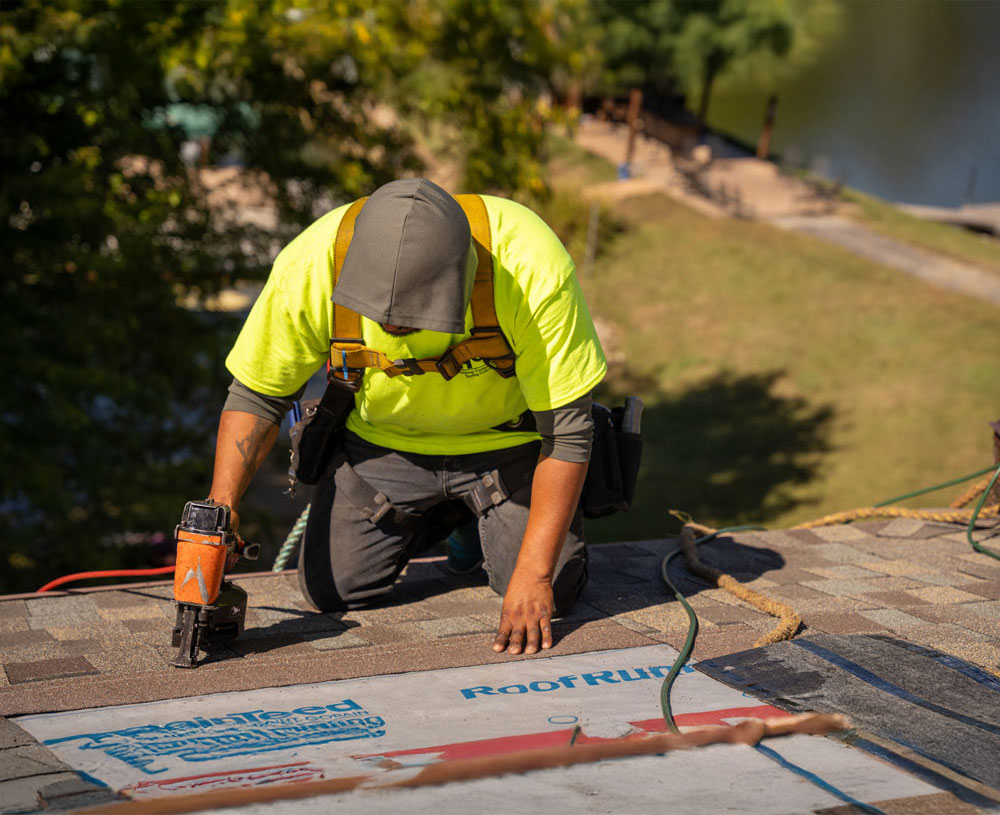 A worker in a yellow safety shirt uses a nail gun while installing shingles on a rooftop, with trees and water visible in the background.