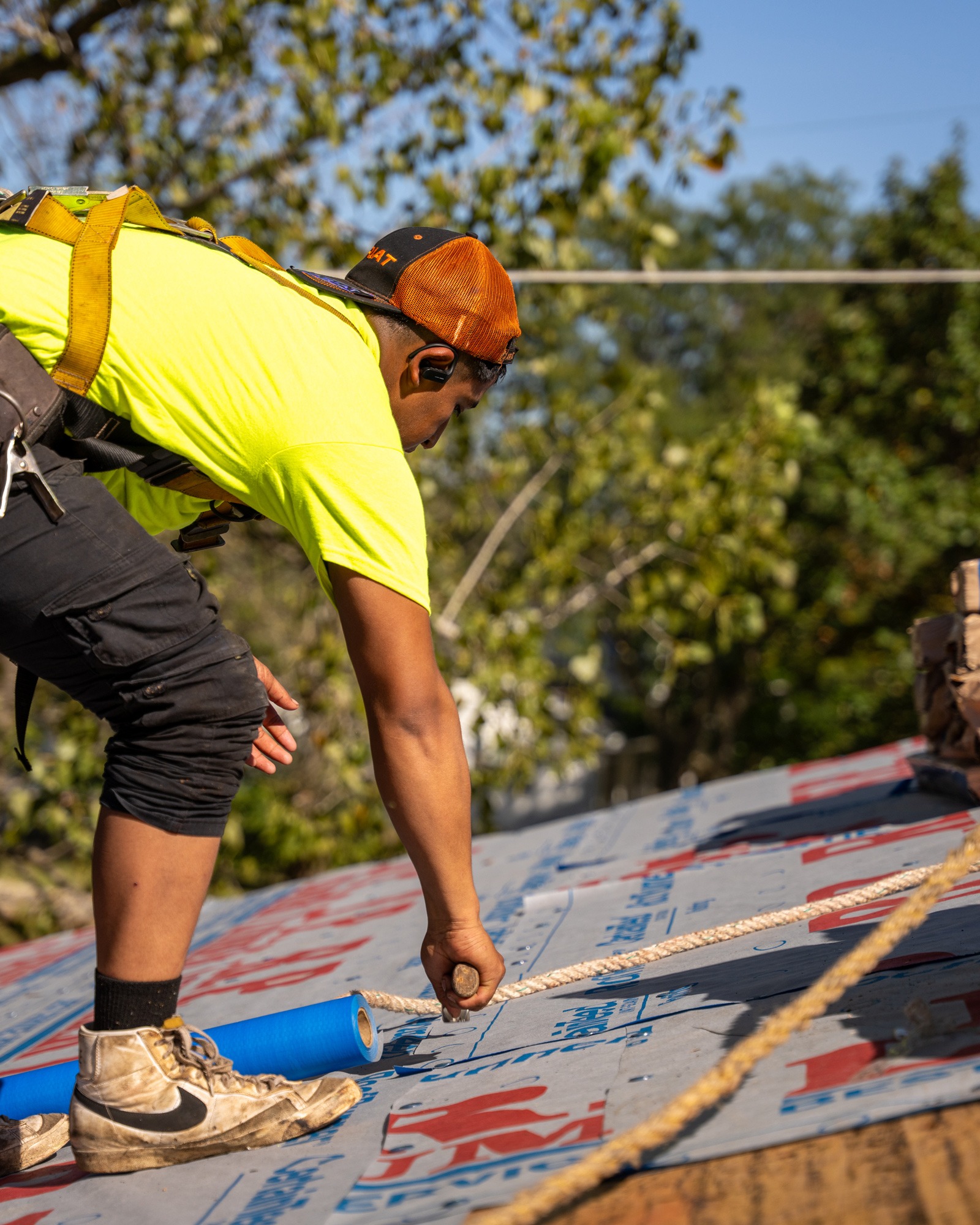 A construction worker in a yellow shirt and safety harness installs roofing material on a house roof, using a tool to secure the material. Trees are visible in the background.