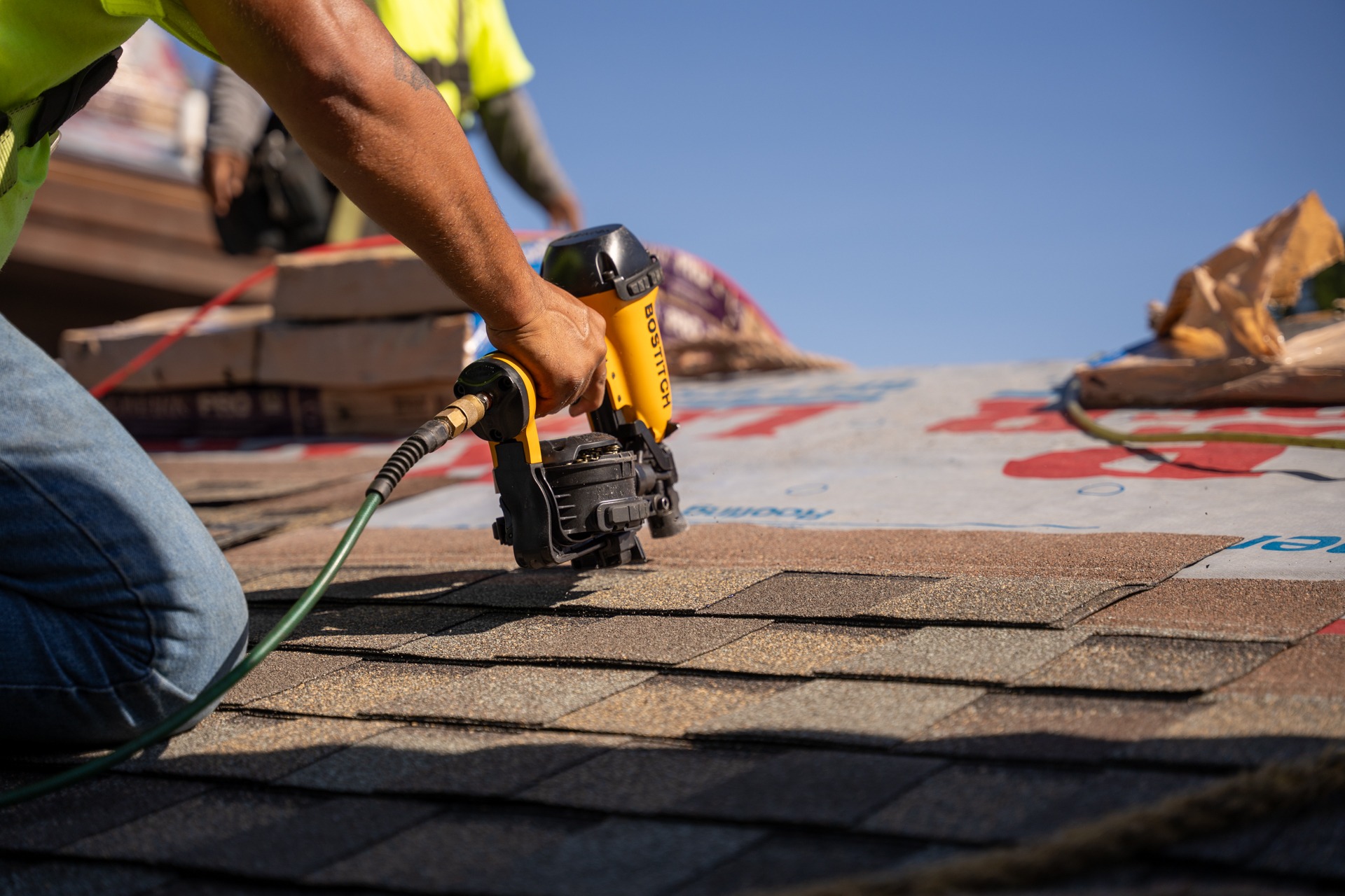 A worker uses a pneumatic nail gun to install shingles on a roof under clear blue sky. A worker uses a pneumatic nail gun to install shingles on a roof under clear blue sky.
