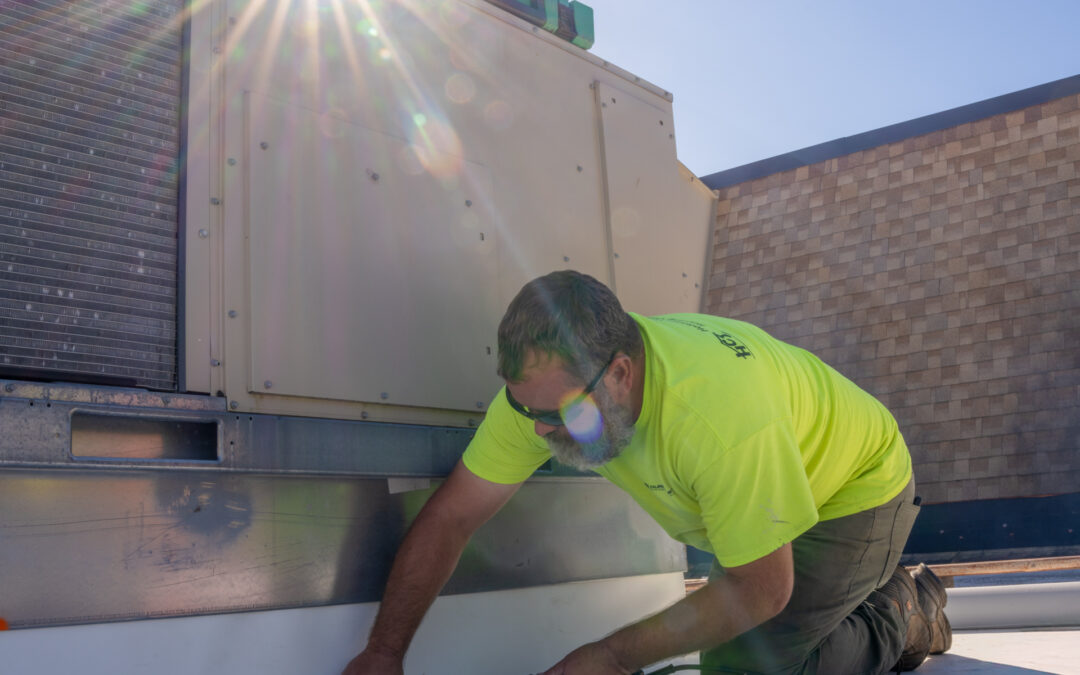 A worker in a neon yellow shirt kneels on a rooftop, sealing the base of an HVAC unit with a tool under bright sunlight.
