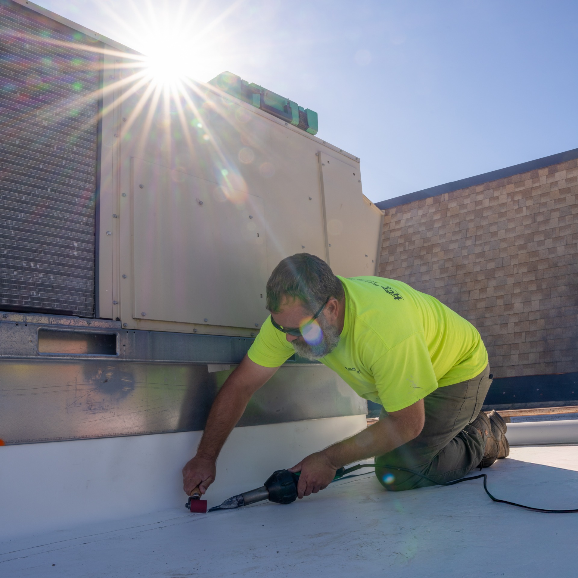 A worker in a neon yellow shirt kneels on a rooftop, sealing the base of an HVAC unit with a tool under bright sunlight. A worker in a neon yellow shirt kneels on a rooftop, sealing the base of an HVAC unit with a tool under bright sunlight.